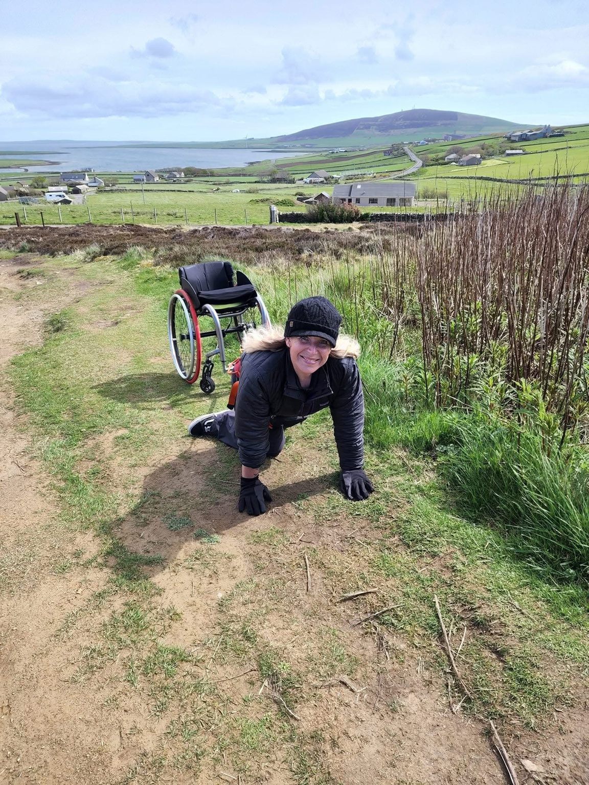 Amanda on hands and knees on a grassy hillside with her wheelchair behind her and the coastline beyond.
