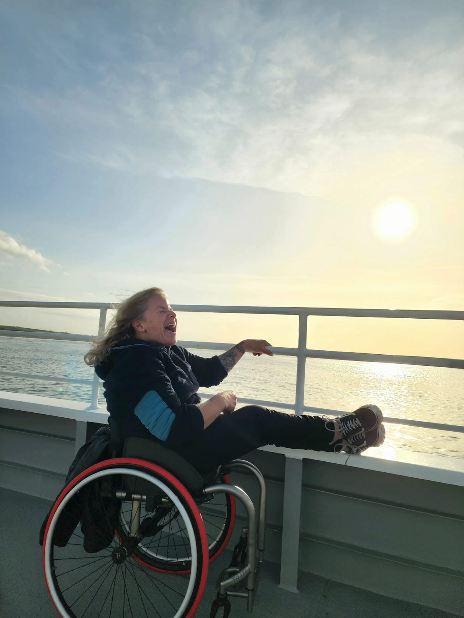 Amanda laughing in her wheelchair on a boat deck during a glowing sunset on the water.