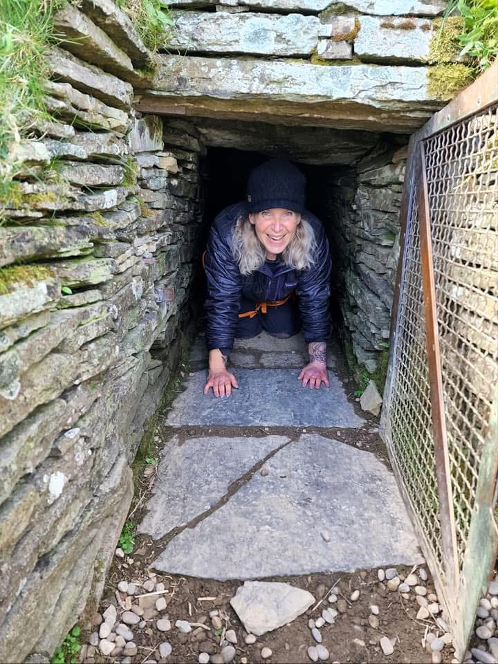 Amanda crawling toward the opening of a low stone passageway, with stacked stone walls and a metal gate.